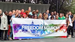 A couple dozen speakers and singers gather on stage behind a rainbow-colored "Freedom March" banner.