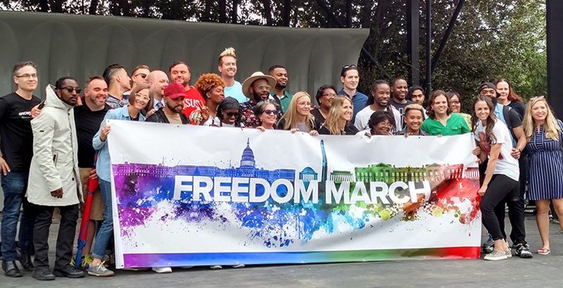 A couple dozen speakers and singers gather on stage behind a rainbow-colored "Freedom March" banner.